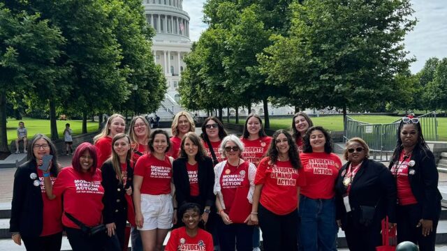 A total of 17 Moms Demand Action and Students Demand Action volunteers, all wearing red branded shirts, pose for a photo during Advocacy Day on Capitol Hill. They stand in front of a staircase with shade trees on either side of the path behind them; the Capitol building is visible between the trees in the background.