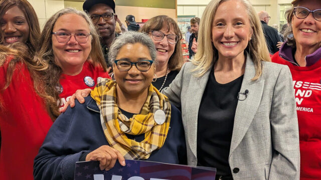 6 Moms Demand Action volunteers stand with Abigail Spanberger, who wears a black shirt, dark pants, and a light grey blazer. One volunteer holds a sign that says 