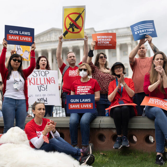 Moms Demand Action volunteers holding up a variety of signs: Gun Laws Save Lives, an assault rifle with a crossed out line through it, and Moms Demand Action and Everytown logo signs