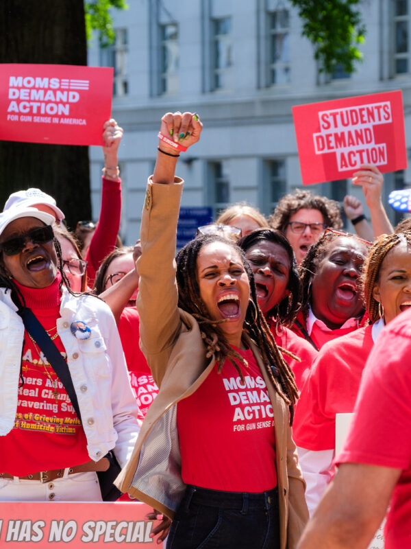 Angela Ferrell-Zabala, executive director of Moms Demand Action and Students Demand Action, stands in the middle of a group of people at a rally. She has her right fist raised and is wearing a tan jacket with a red Moms Demand Action t-shirt. She is framed by a red Moms Demand Action sign raised on the left of the image and a red Students Demand Action sign raised on the right of the image.