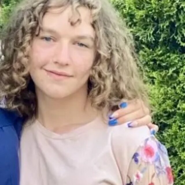 Pauly Likens, a white trans girl, smiles for a photo in front of a green foliage background. Pauly's hair is light and curly; she has bangs, and the rest of it is loose and curls just above her shoulders. She wears a blush pink long-sleeve top that has mesh and floral details on the sleeves.