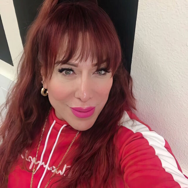 Reyna Hernandez, a Latina trans woman, poses for a selfie; her lips are slightly pursed. She wears her long wavy red hair in a high ponytail and has bangs that frame her face. She is wearing a red sweatshirt with a single white vertical stripe on the sleeve; she also wears hot pink lipstick.