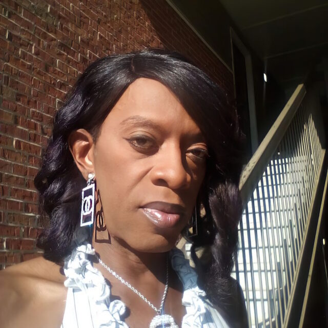San Coleman, a Black gender non-conforming person, takes a selfie. San is standing outside of brick building that has a staircase. San wears a white striped halter top, a silver necklace with a large metal pendant, and large silver earrings. San's shoulder-length dark hair is parted on the side and is curled.