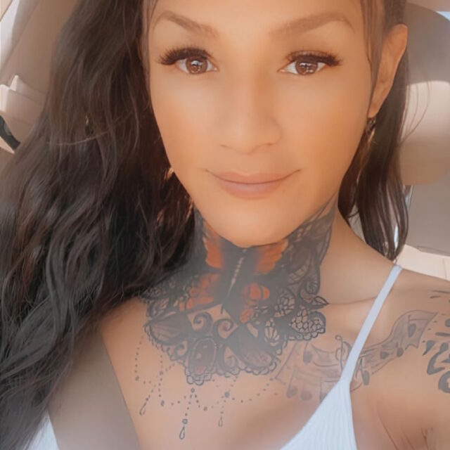 Sasha Williams, a multiracial trans woman, gives a soft smile to the camera while taking a selfie. Her long dark hair is wavy and pulled into a high ponytail with a rainbow leopard-print scrunchy. She is wearing a white tank top and has a lacy neck tattoo covering her throat and her upper chest.