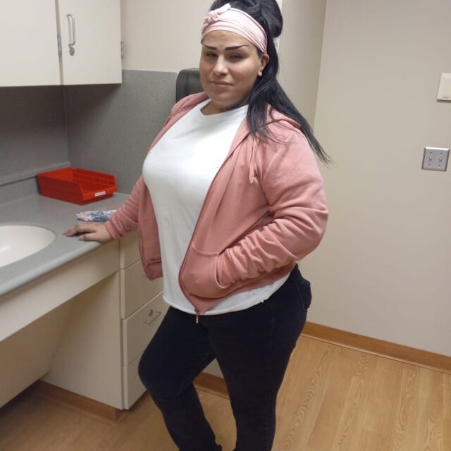 Savannah Ryan Williams, a Cuban and Native trans woman, poses for a photo. She wears black pants, a white t-shirt, and a blush-pink unzippered hoodie over the t-shirt. Her long black hair is pulled back with a pink tie-wrap headband.