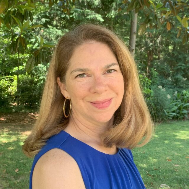A woman with light skin and shoulder-length, light-brown hair is wearing gold hoop earrings and a cobalt blue, sleeveless blouse. She is softly smiling at the camera and is standing in what appears to be a backyard, with green trees and green grass visible in the background. She is pictured from the shoulders up; her torso is turned about 80 degrees away from the camera, and her head is directly facing the camera.
