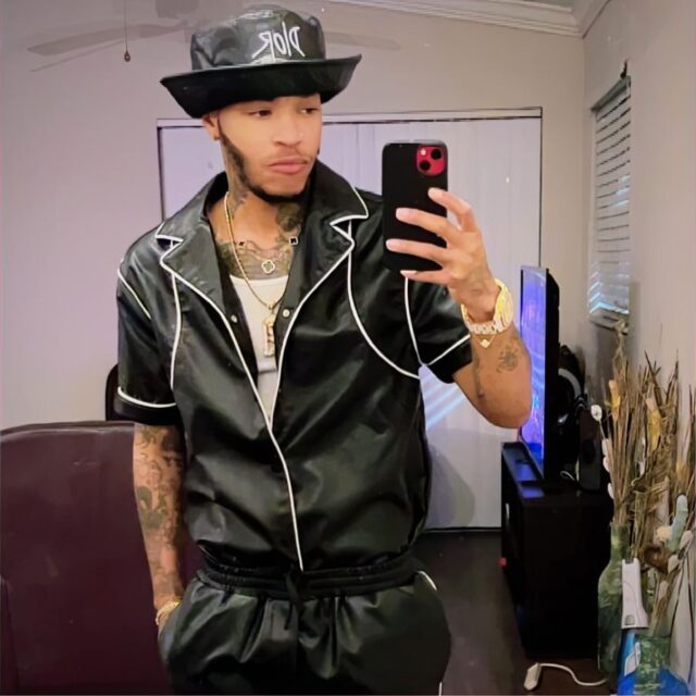 Tee Arnold, a Black trans man, wears a black hat with an oversized brim, a black leather short-sleeve shirt, a white tank top, and black bottoms. He has a white watch on his left wrist and is taking a selfie in the mirror with his phone, which he holds in his left hand. His right hand is in his pocket.