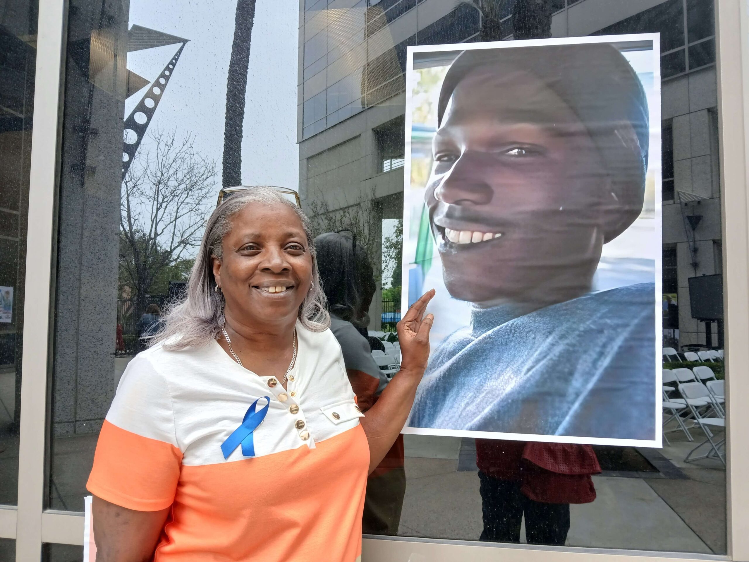 Giesela Tanner, a Black woman with grey hair, stands next to a photo of her son, Ellis. Ellis was shot and killed; his killing remains unsolved. Giesela is wearing a block-color t-shirt with white on the top third of the shirt and orange on the bottom two thirds; she has a blue ribbon pinned to her shirt. Ellis is wearing a dark beanie and a light sweatshirt in the photo; he is smiling at the camera.