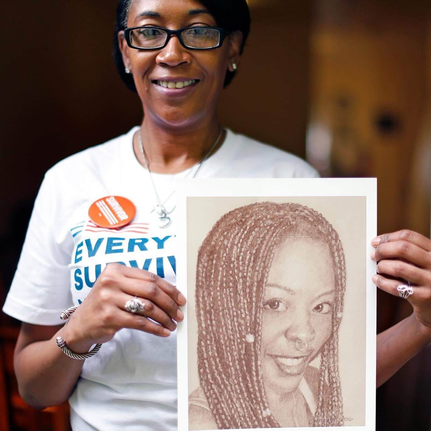 Nicole Gardner, a Black woman, holds a pencil portrait of her daughter, Ronique. Ronqiue was shot and killed; her killing remains unsolved. Nicole is wearing a white Everytown Survivor Network t-shirt, an orange Survivor pin, and black rectangular-frame glasses. In the pencil sketch, Ronique is smiling at the viewer; her long hair is parted down the middle and braided with beads; it falls below her shoulders.