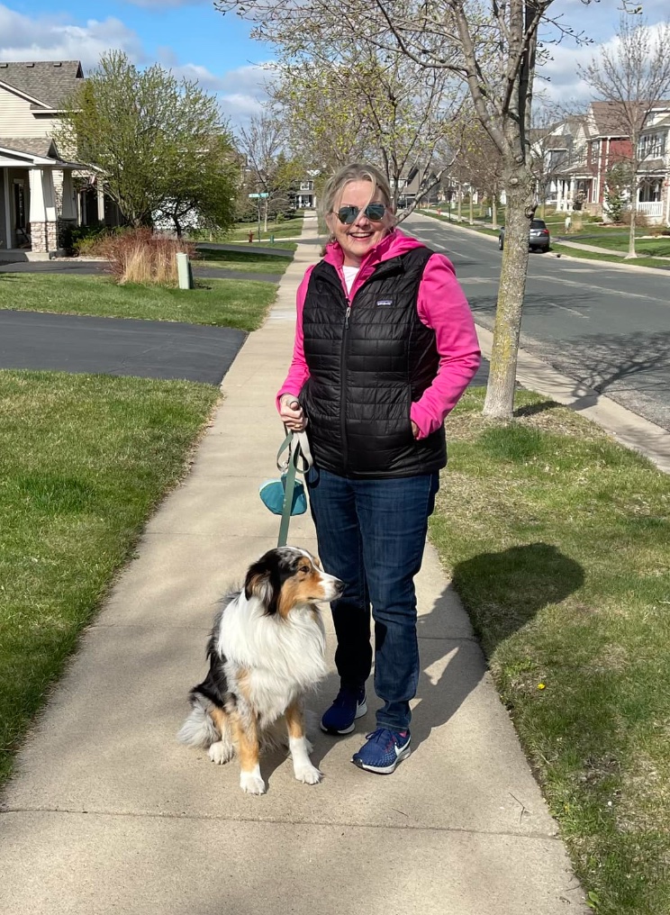 Patty Matthews wears dark jeans and a black puffy vest layered over hot pink zip-up jacket. Patty is holding a leash in her right hand; the border collie she is walking sits on the sidewalk next to her. 