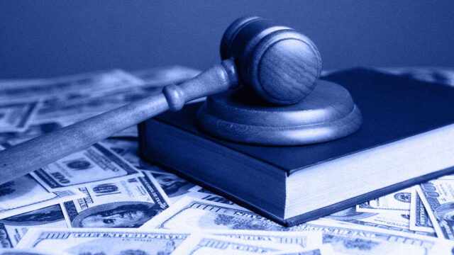 A courtroom gavel rests on top of a book. Underneath both, hundred-dollar bills are scattered on a table. The image represents the financial investment in the 2025 Wisconsin Supreme Court Election.