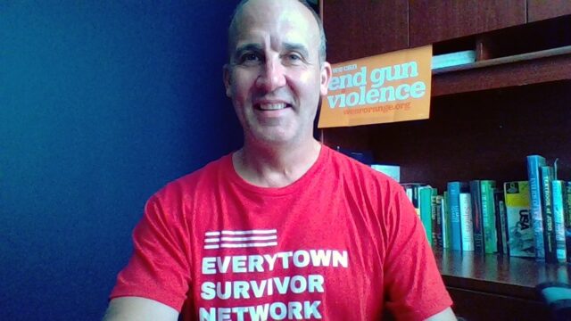 James smiles while wearing his Everytown Survivor Network shirt. Behind him is a sign that reads We Can End Gun Violence.