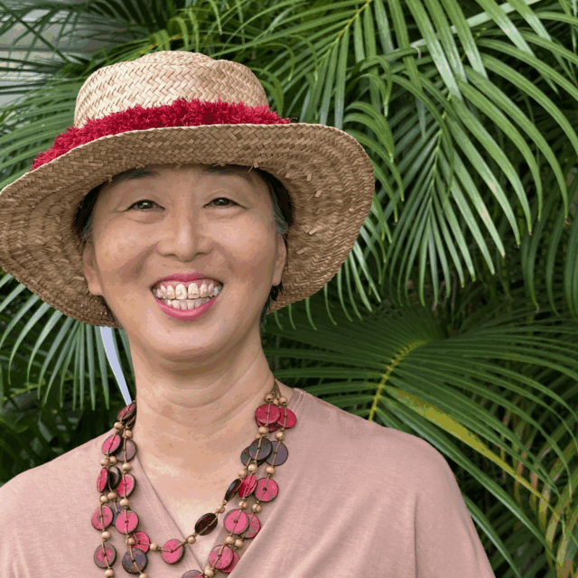 A photo of Elna Nagasako from the waist up, who is pictured in front of some ferns while wearing a dusky rose-colored pink wrap top, a chunky multi-strand necklace, and a straw hat.